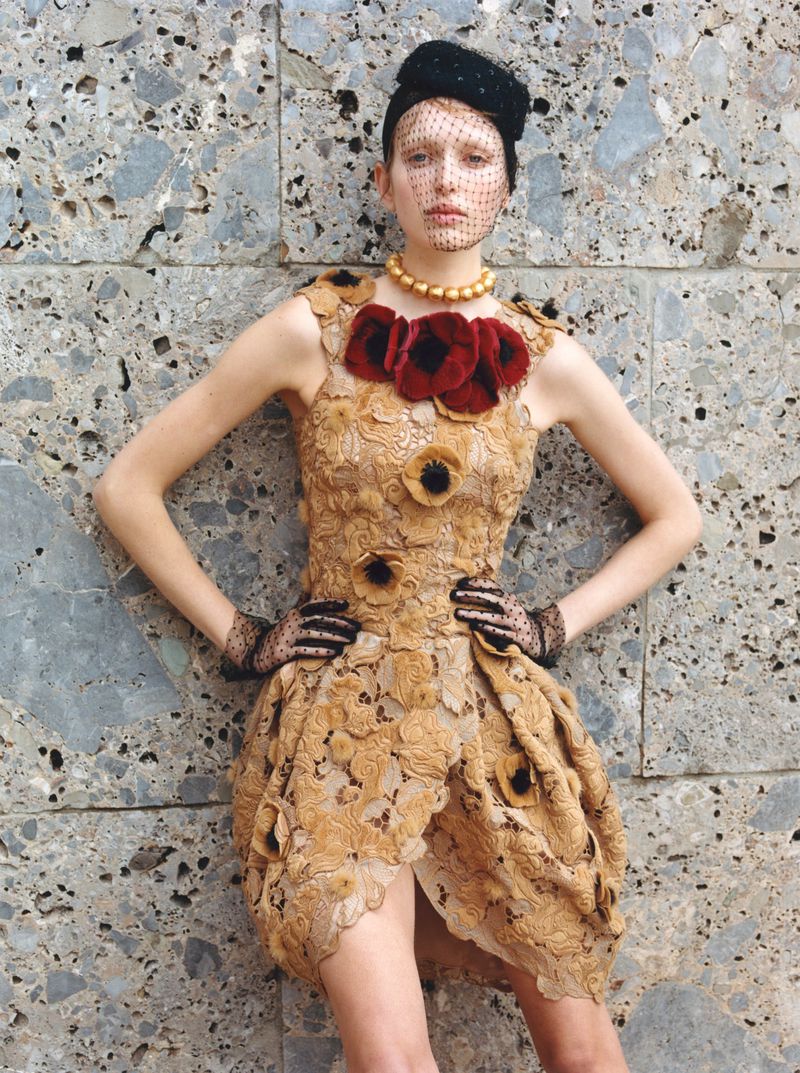 A woman poses in a tan, floral-patterned dress and black netted headpiece against a grey-patterned wall in an eye-level, medium shot.
