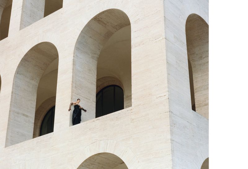 A woman in a black dress stands framed in an archway of a stone building with an array of archways in its architectural design.