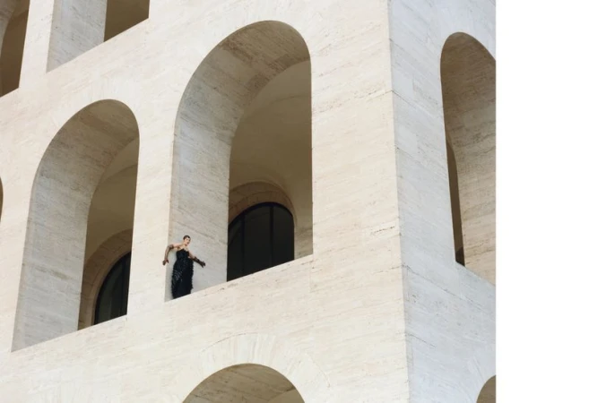 A woman in a black dress stands framed in an archway of a stone building with an array of archways in its architectural design.