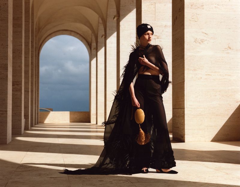 A woman in a sheer black outfit poses in a colonnade with dramatic lighting, wearing a feathered cape and holding a gold tassel purse.