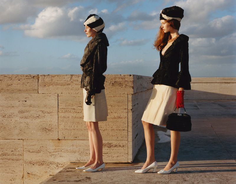 Two fashion models in vintage-inspired outfits featuring textured jackets, hats, gloves and purses in an outdoor setting with light sky.