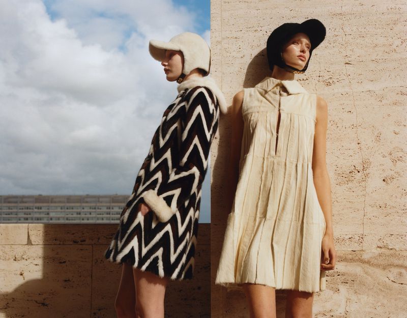 Two women pose in fashionable coats and hats; one in white and black, the other in cream, with a textured background.