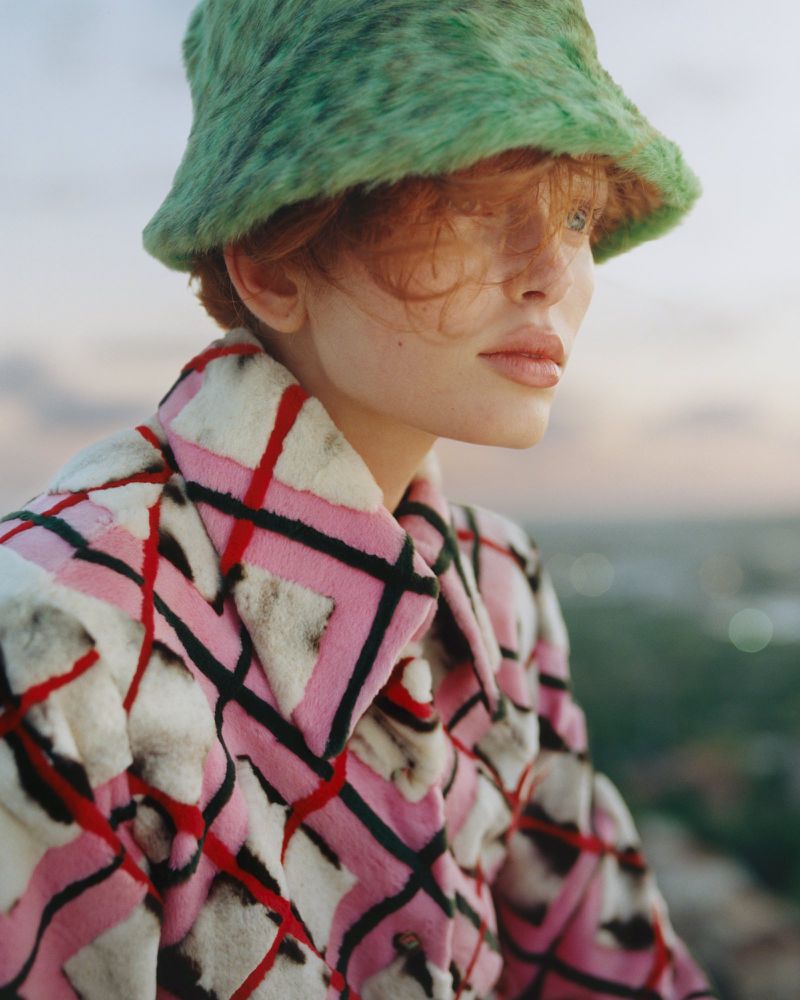 Close-up of a redhead woman wearing a fuzzy green bucket hat and patterned jacket in an outdoor setting with muted colors and diffused lighting.