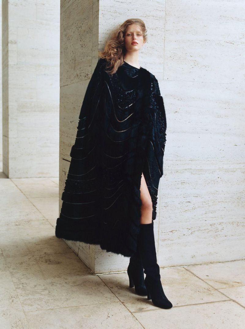 A lone fashion model in a dark cape and boots poses in a textured urban environment. Natural light highlights a striking, monochromatic look.