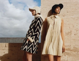 Two female models posing outdoors wearing patterned coats and hats against stone architecture and a cloudy sky.