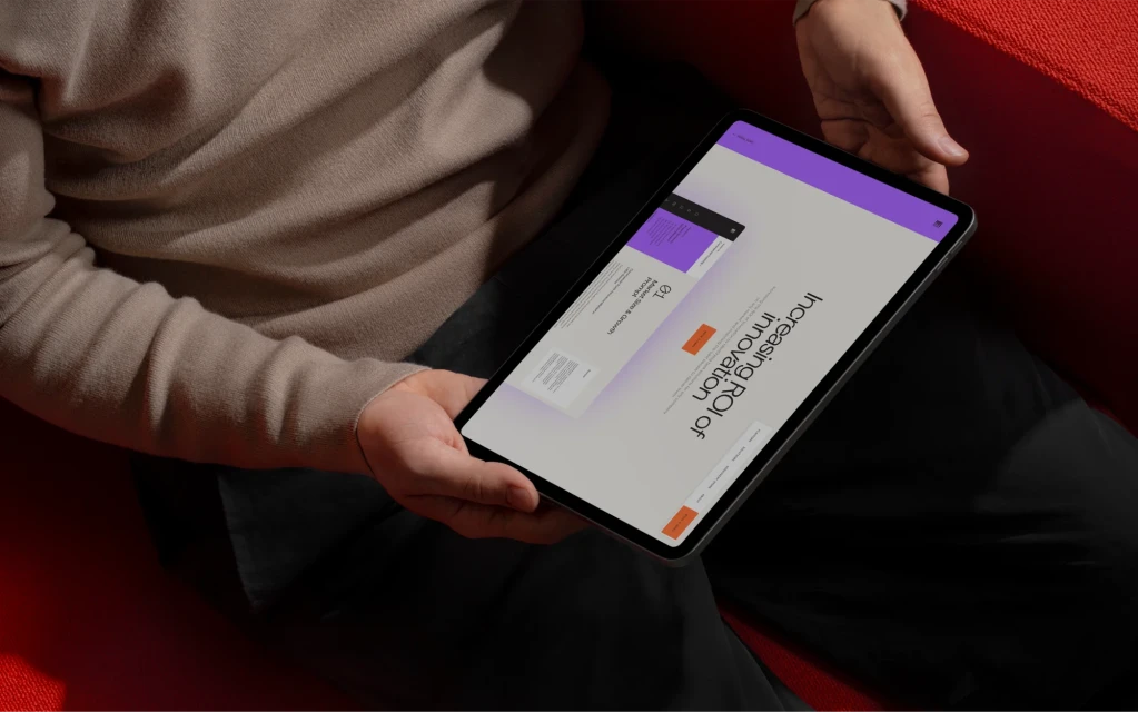 Adult holding a tablet displaying 'Increasing ROI of Innovation'. The scene is indoors, with a red seat and neutral-toned sweater.