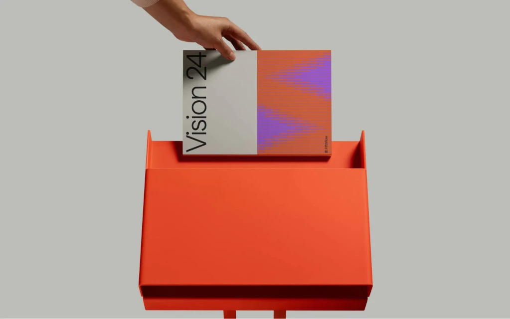A magazine called Vision 24 is being put into a bright orange metal magazine rack in a studio setting with a light grey background.