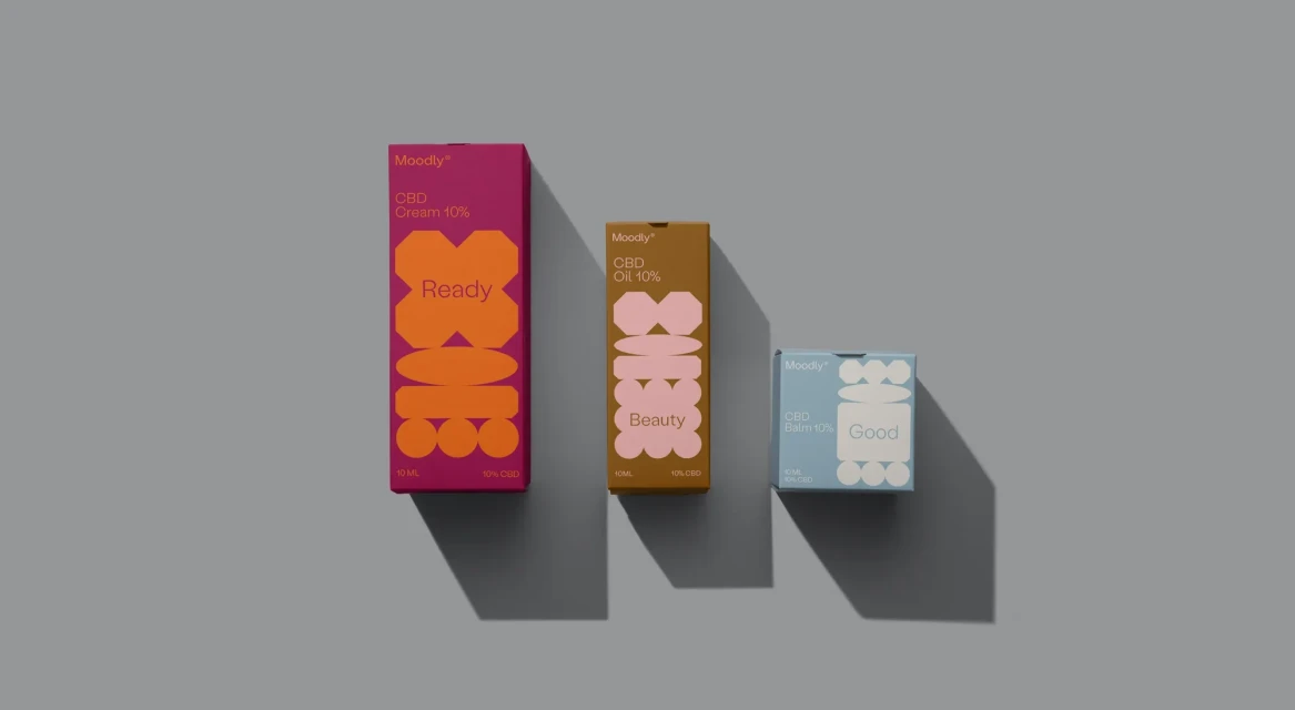 Three boxes of Moodly CBD products, including cream, oil, and balm, are arranged on a grey surface in a minimalist composition.