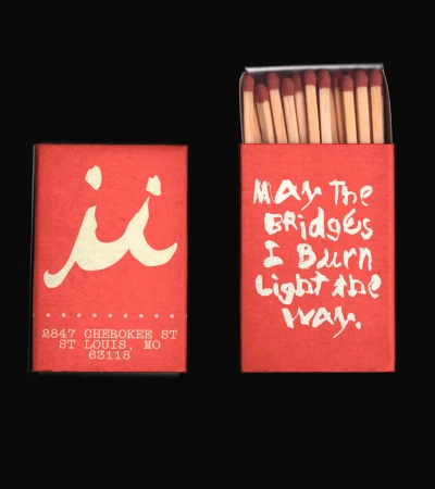 Two red matchboxes. One has matches; both have cream-colored text including an address and the saying, "May the bridges I burn light the way."