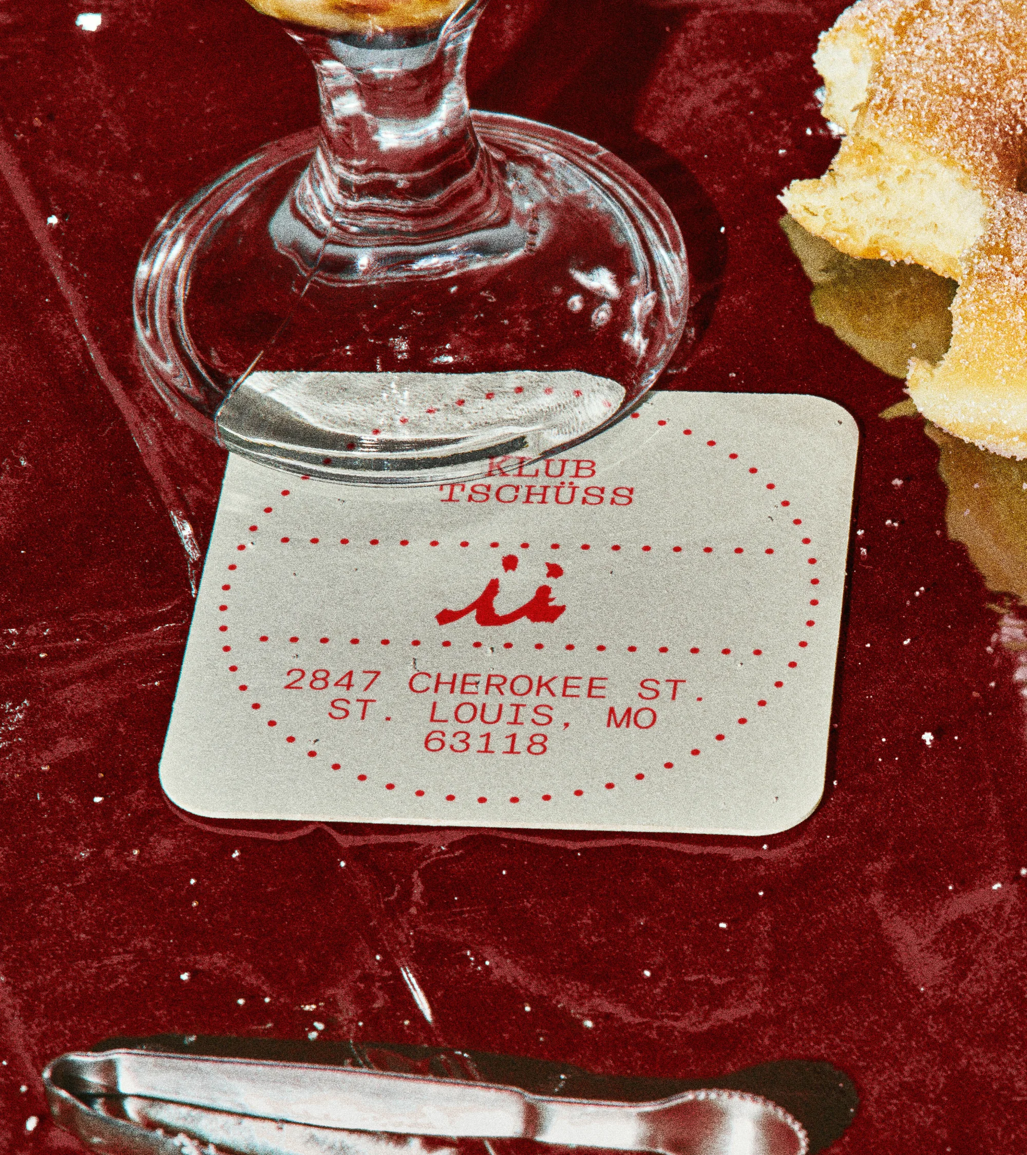 Close-up shot of a cocktail glass and a business card for Klub Tschuss on a red surface with tongs, suggesting a bar or restaurant setting.