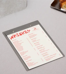 A metal clipboard holding a whiskey menu resting on a light grey textured table with a pastry tray nearby