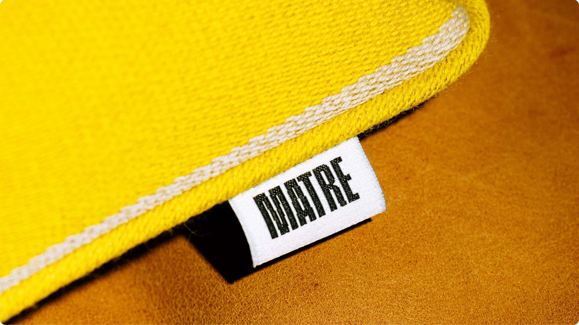 Close-up of yellow fabric with white stripe and a tag with the name 'MAITRE' on a textured brown surface. Focus on fabric texture.