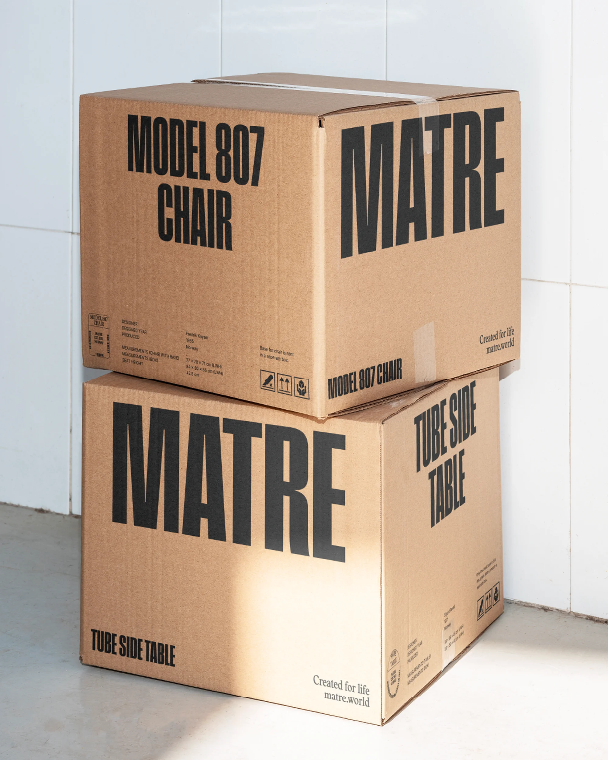 Stacked cardboard boxes labeled "MATRE" display "Model 807 Chair" and "Tube Side Table" on a white tiled floor.