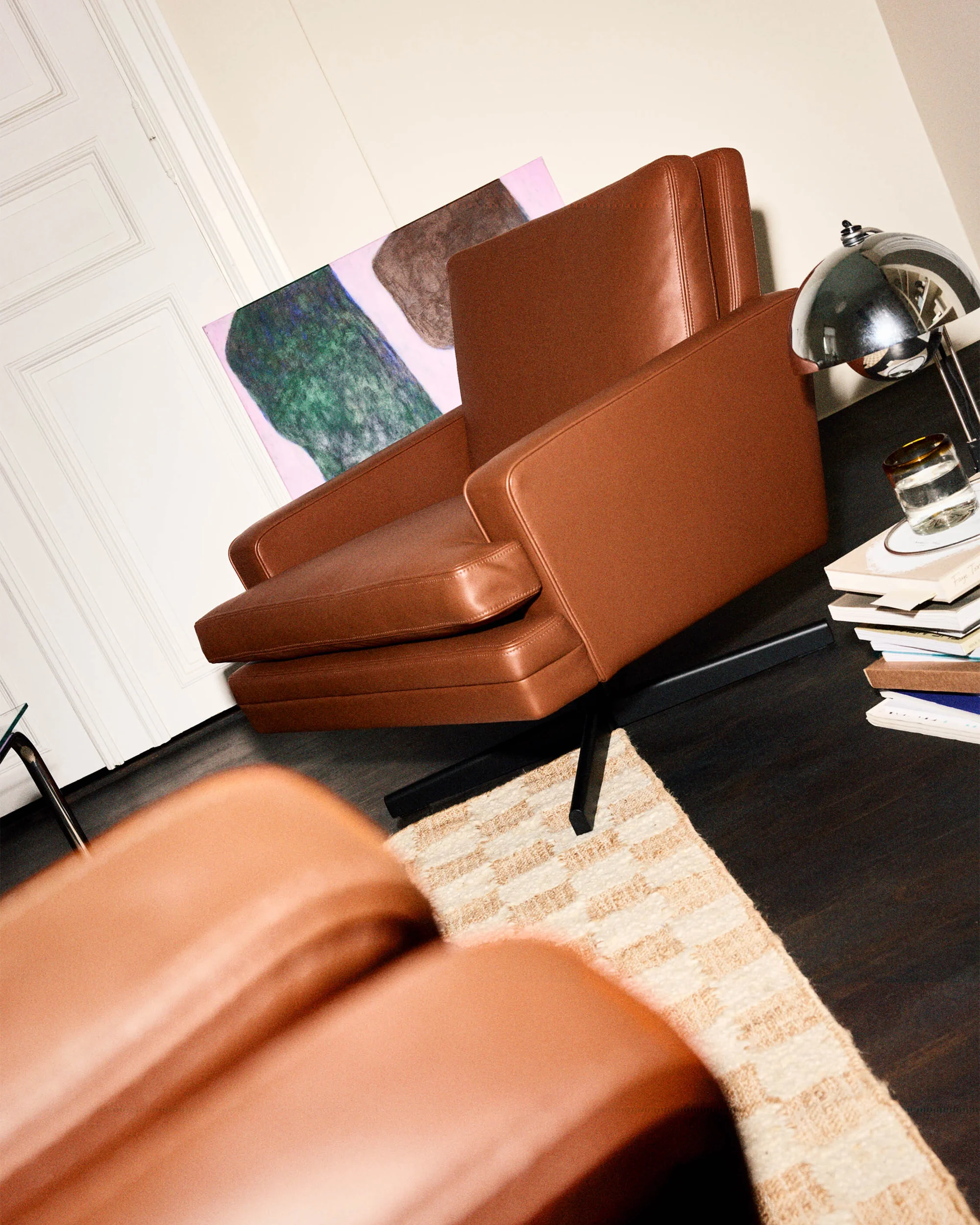 Modern interior features tan leather armchair, abstract art, and table lamp on dark wood floor with a patterned rug. Interior design.
