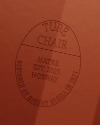 Embossed oval typographic label reading Tube Chair Matre Est. 2023 Norway on a textured terracotta surface.