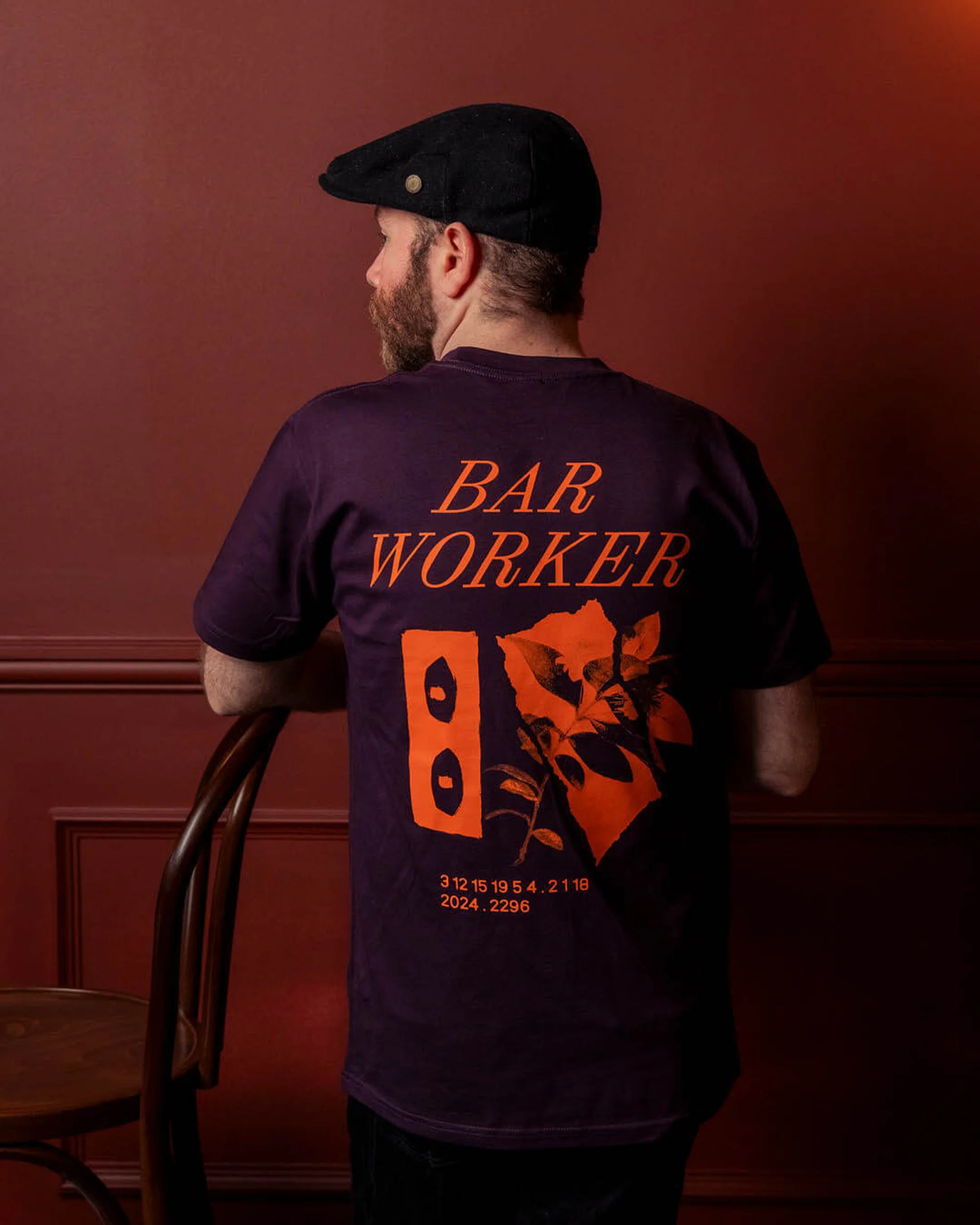 Adult male wearing a purple t-shirt with 'Bar Worker' graphic, standing indoors with his back to the viewer, next to a chair.
