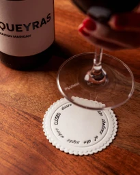 Close-up of a wine glass held above a white scalloped coaster with text, next to a wine bottle on a wooden surface.
