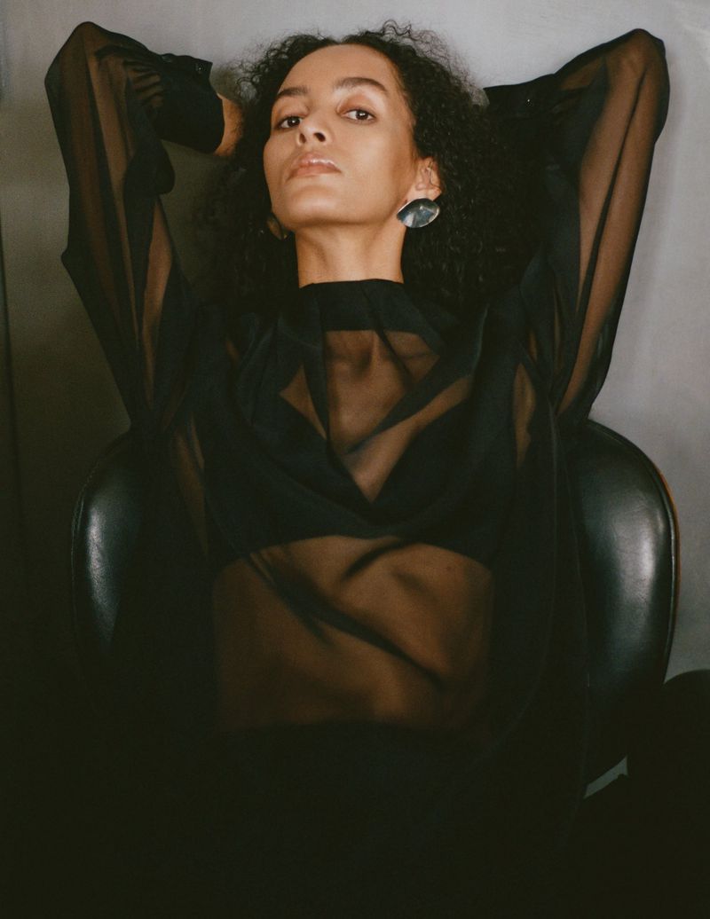 Woman in sheer top poses in black chair against grey wall. Portrait of a young woman indoors. Fashion editorial shot.