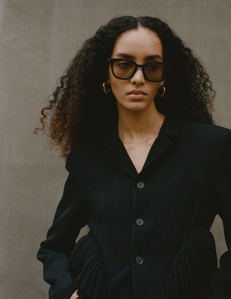 Portrait of a woman with curly hair wearing sunglasses and a black jacket against a neutral backdrop, exuding a stylish and modern aesthetic.