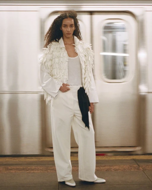 An adult woman with curly hair poses in a white outfit with a fringed jacket against the blurred backdrop of a subway train.