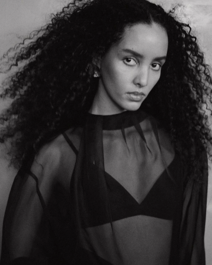 Portrait of a woman with voluminous dark curly hair wearing a sheer black top and bra against a textured studio background.