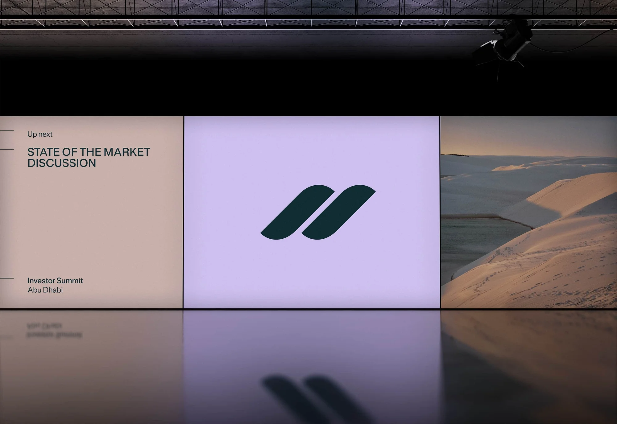 Presentation slide with geometric logo, informational text, and a landscape view of desert sand dunes at dusk, reflected in a glossy floor.