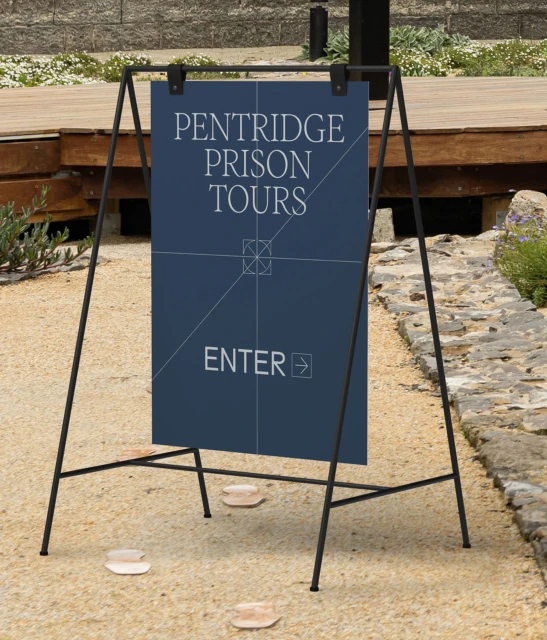 Sign for Pentridge Prison Tours stands outdoors on gravel with a wooden platform in the background, advertising prison tours.