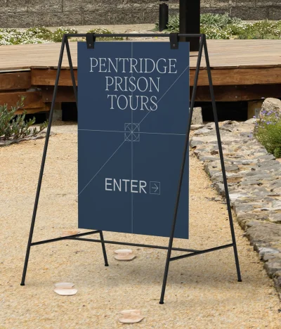 A-frame dark blue sign with white lettering reading Pentridge Prison Tours and ENTER on a gravel path outdoors.