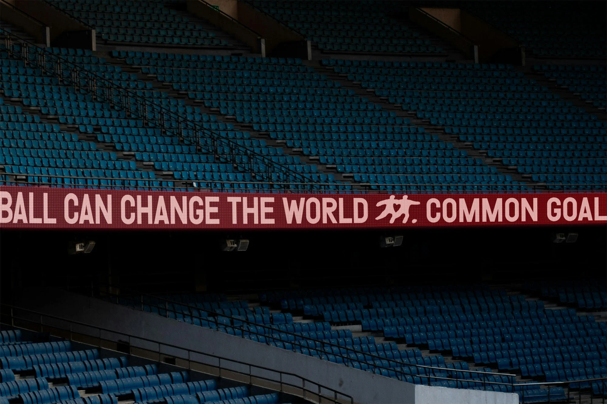 Empty stadium seating with an LED sign reading, 'FOOTBALL CAN CHANGE THE WORLD' and 'COMMON GOAL' in white text against a red background.