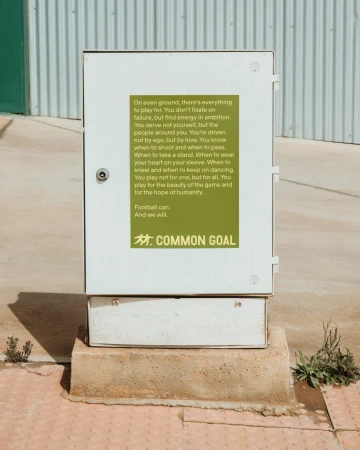 Outdoor shot of a white electrical box with an inspirational green poster that reads "COMMON GOAL" on a gray and tan sidewalk.