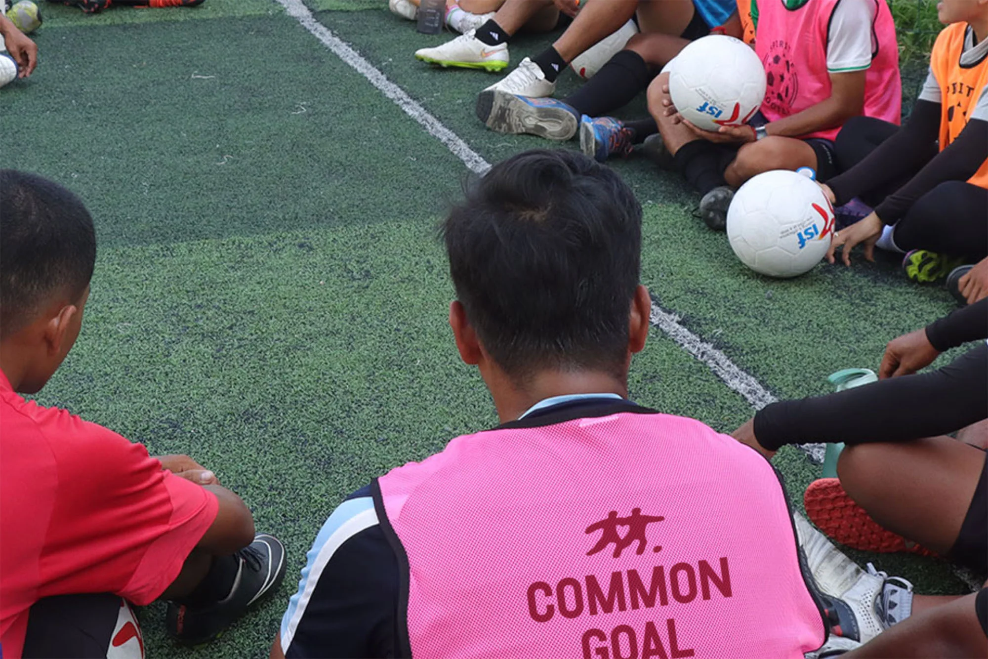 Group of people sit on the ground in a circle formation on an artificial turf soccer field. A man wears a pink vest with the words 'Common Goal'.