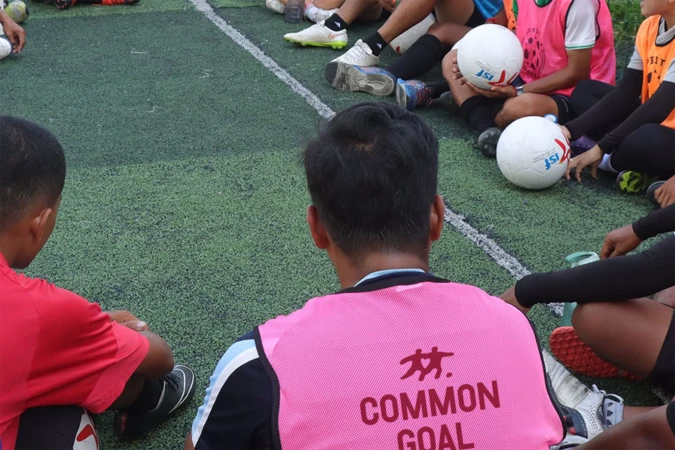Group of people sit on the ground in a circle formation on an artificial turf soccer field. A man wears a pink vest with the words 'Common Goal'.