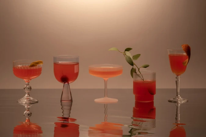 Five stemmed cocktail glasses filled with vibrant red and peach colored cocktails, garnished with citrus peels and green leaves, reflecting light.