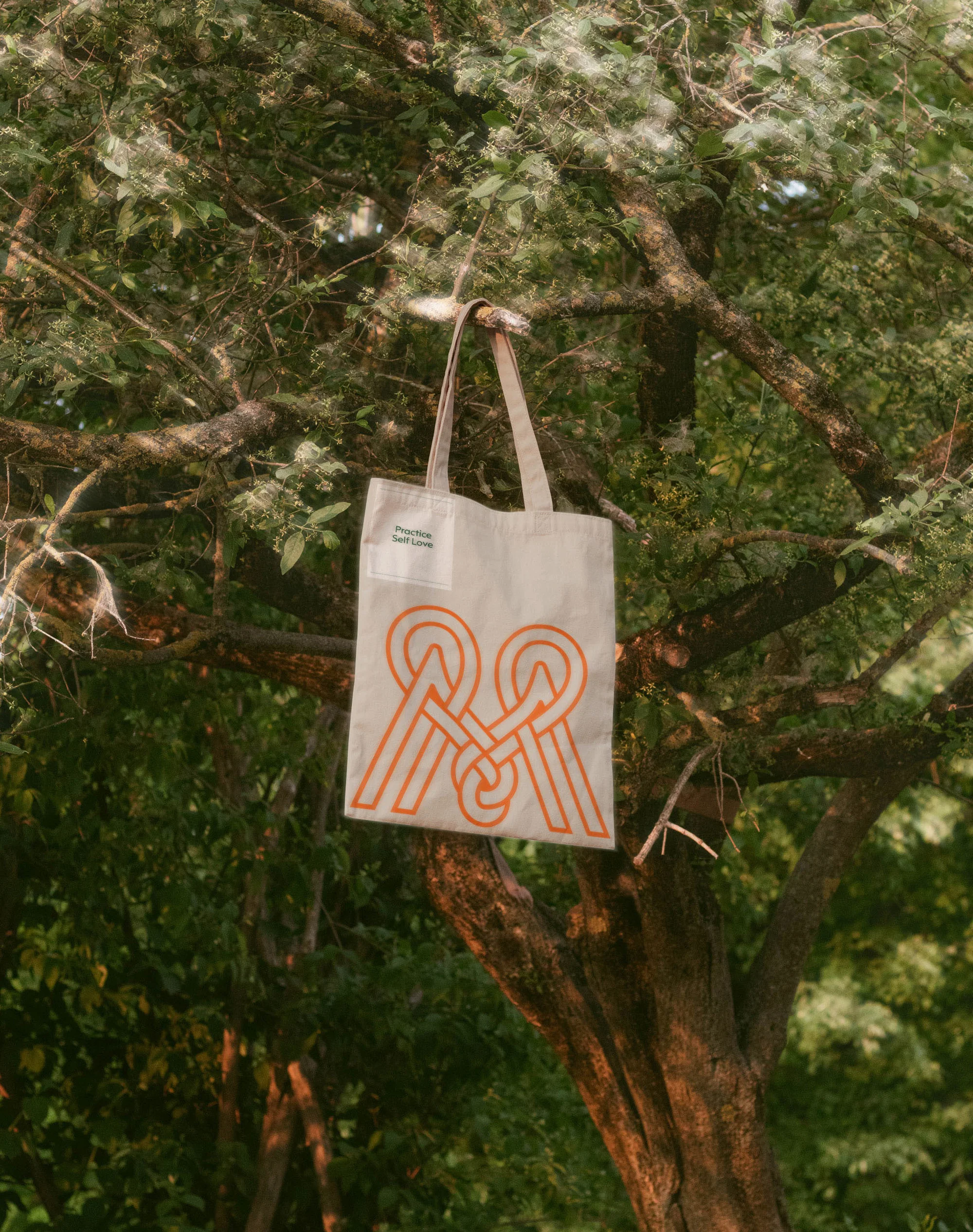 Tote bag with orange logo hanging from a tree branch surrounded by green leaves and branches in natural daylight conditions.