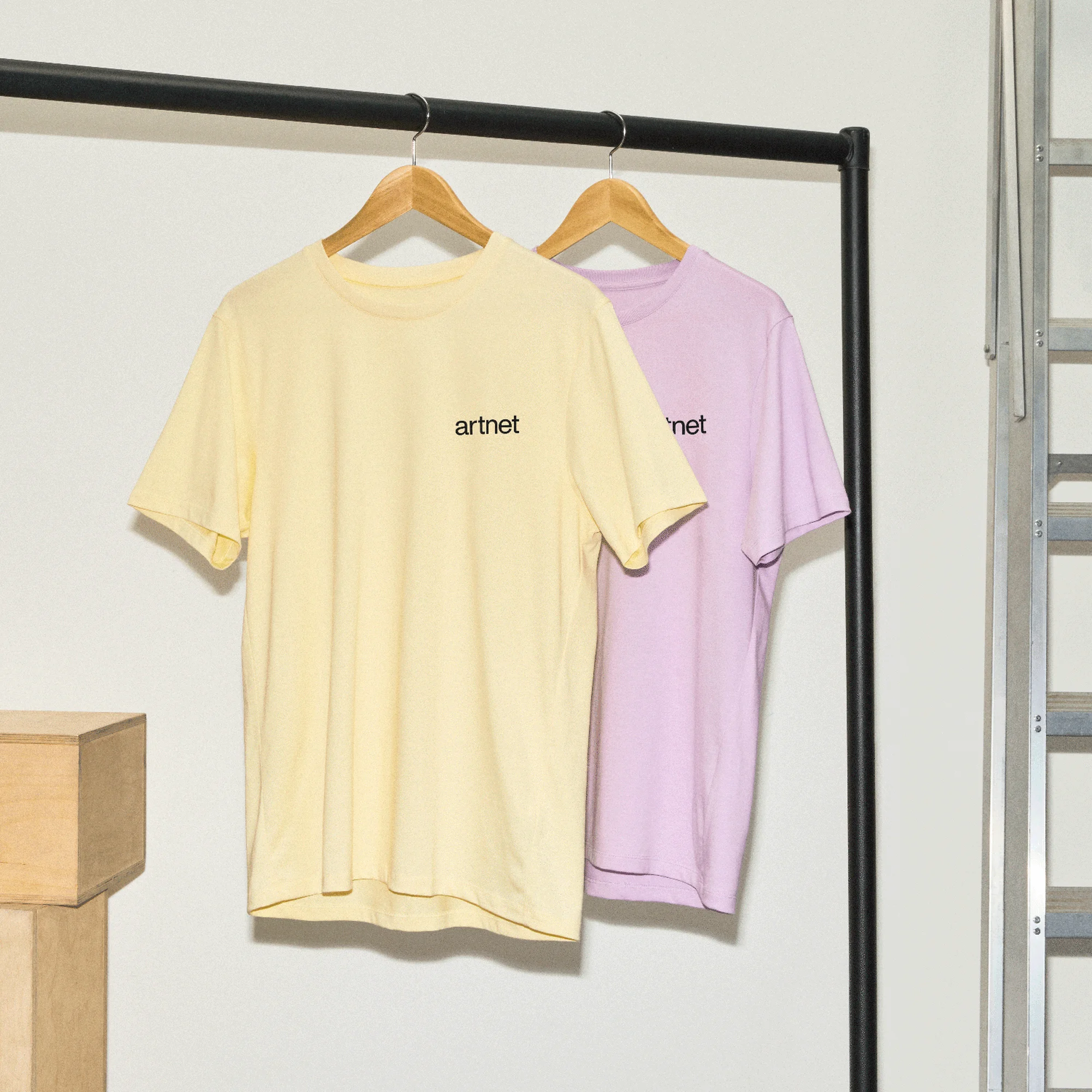 Two pastel t-shirts with logo hanging on rack, one yellow and one light purple, in a minimalist indoor fashion studio setting.