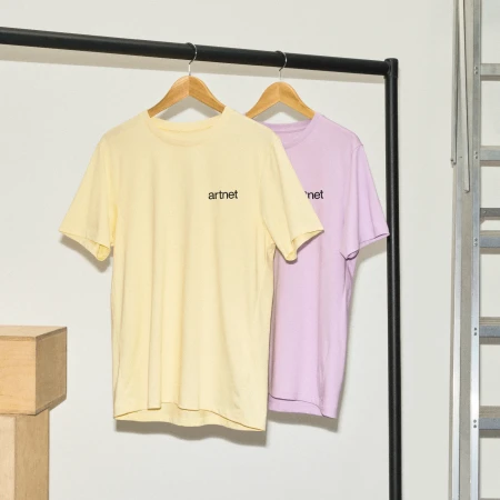 Two pastel t-shirts with logo hanging on rack, one yellow and one light purple, in a minimalist indoor fashion studio setting.