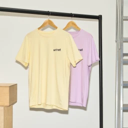 Two t-shirts in yellow and light purple hanging on wooden hangers from a black rack against a white wall.