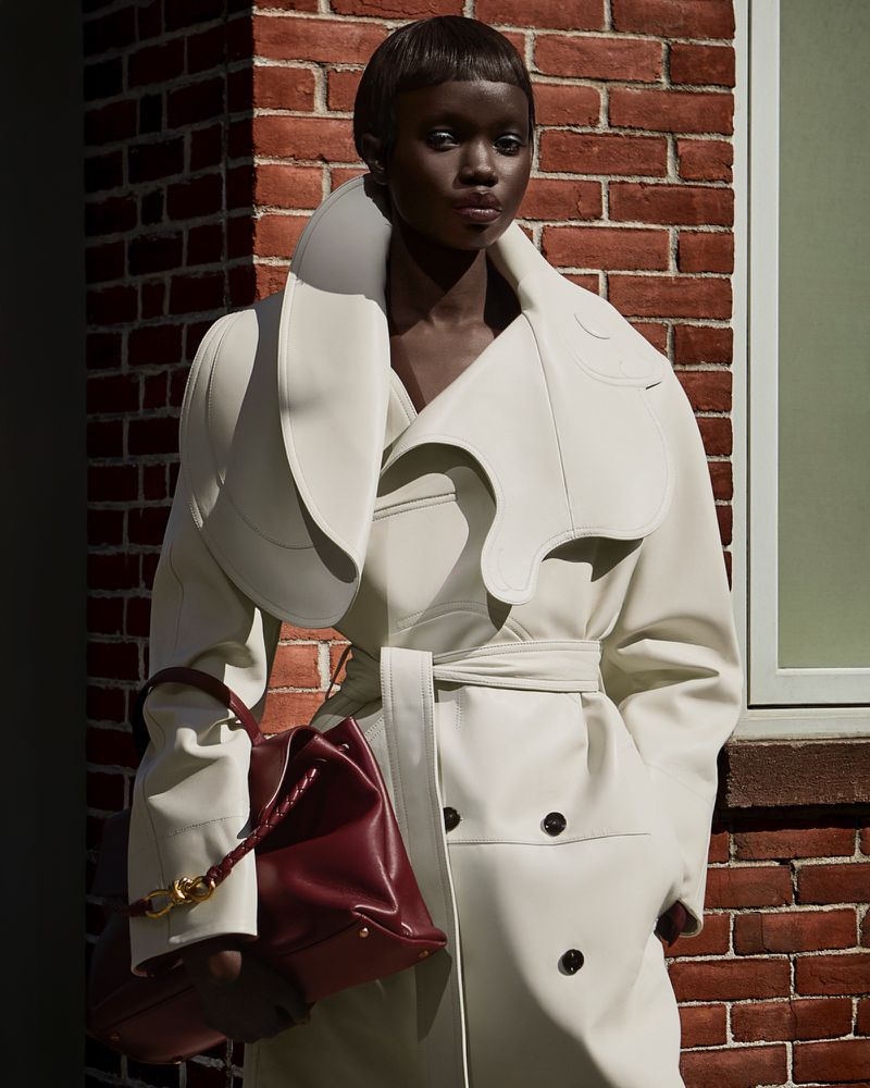 Fashion editorial shot of a woman in an off-white, belted trench coat with a burgundy leather bag against a brick wall.
