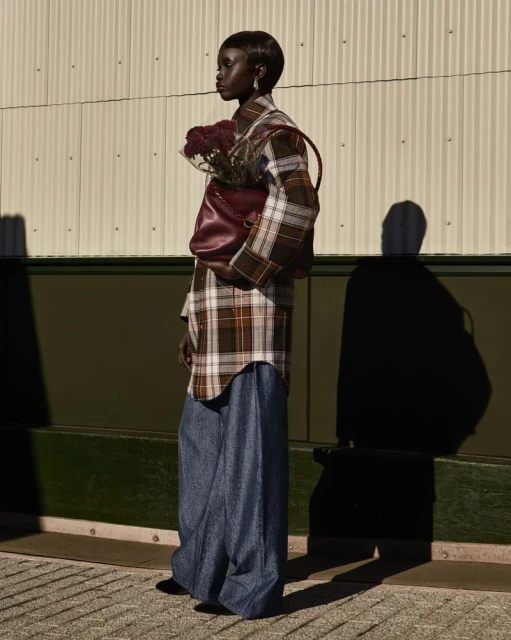 A tall, slim, dark-skinned model poses in an outdoor setting, wearing a plaid coat, wide blue trousers, and carrying a maroon leather handbag.