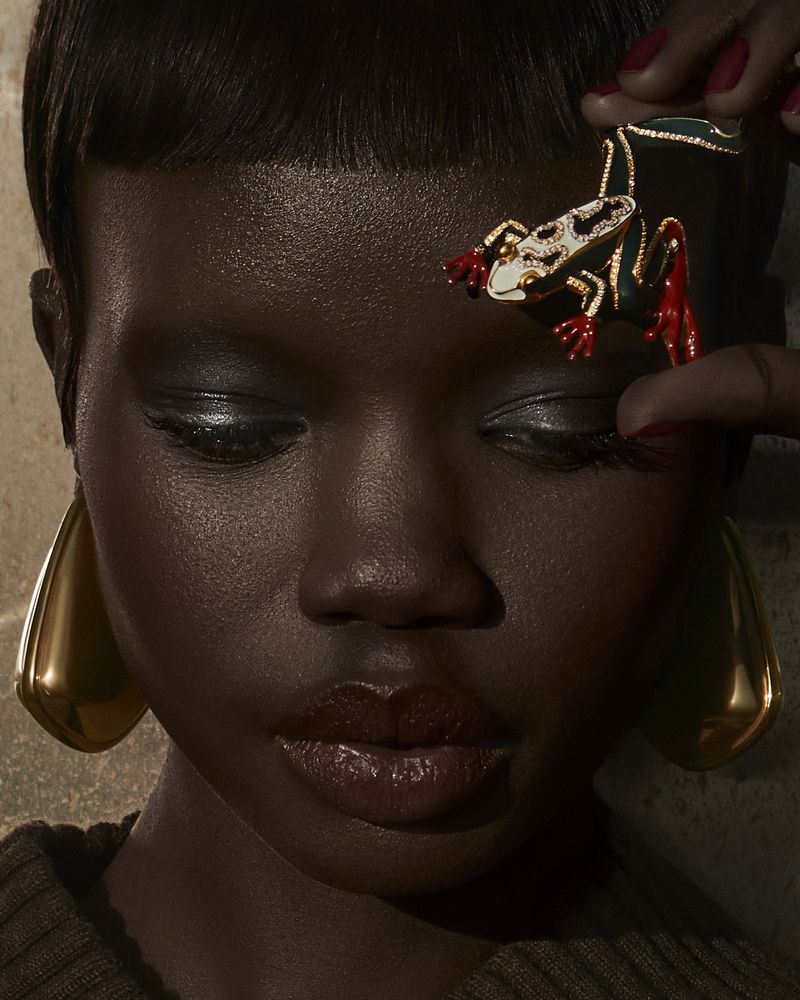 Close-up of a dark-skinned woman with dramatic eye makeup and large gold teardrop earrings, posing with a frog-shaped jewelry piece.