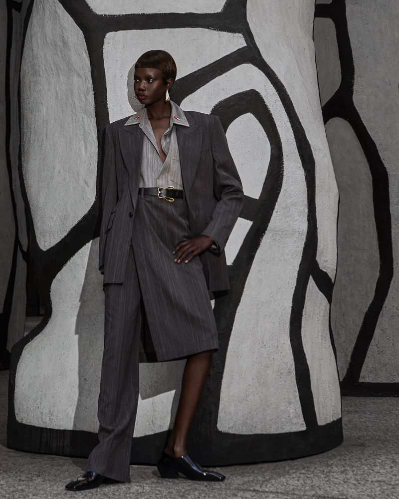 Fashionable adult Black woman poses in a pinstripe suit, collared shirt, and black loafers against an abstract black and white backdrop.