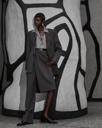 Fashionable adult Black woman poses in a pinstripe suit, collared shirt, and black loafers against an abstract black and white backdrop.