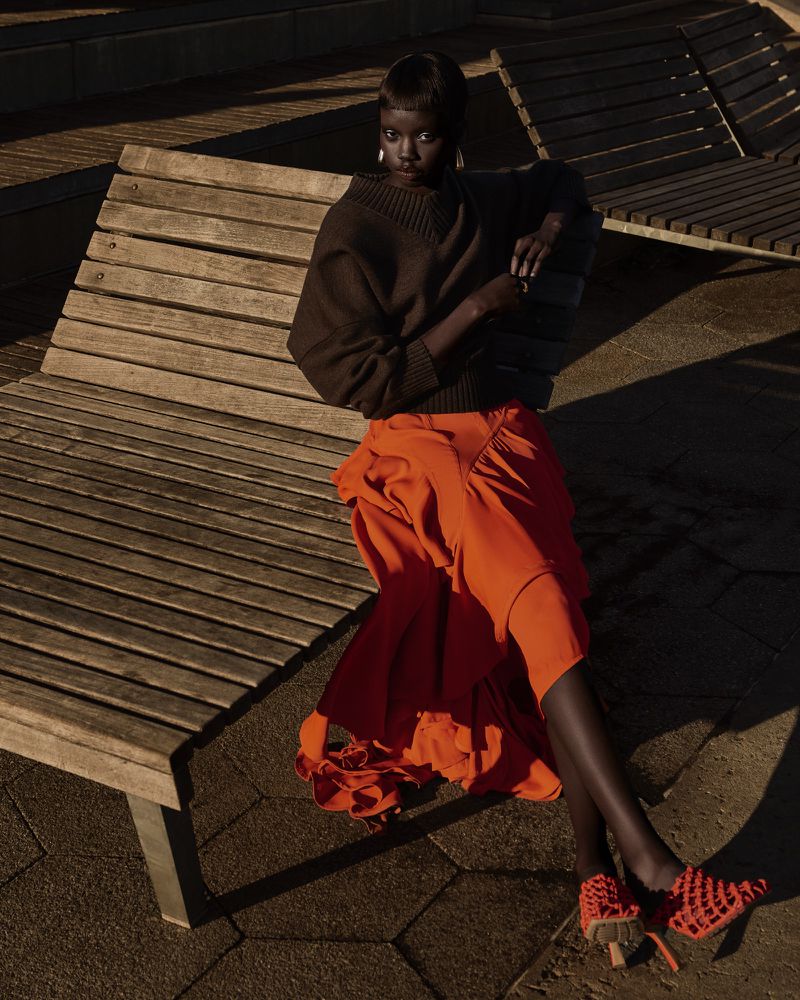 A fashionable Black woman poses on a wooden park bench in a brown sweater and orange skirt with matching shoes in outdoor lighting.