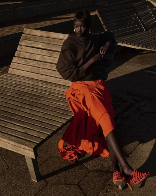 A fashionable Black woman poses on a wooden park bench in a brown sweater and orange skirt with matching shoes in outdoor lighting.