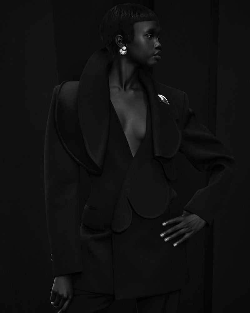 Monochrome studio portrait of a Black female model with short hair wearing a tailored jacket and earrings against a dark backdrop.