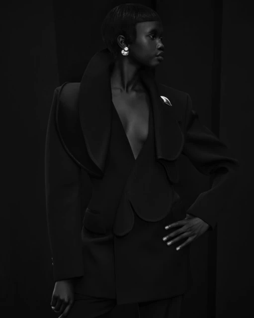 Monochrome studio portrait of a Black female model with short hair wearing a tailored jacket and earrings against a dark backdrop.