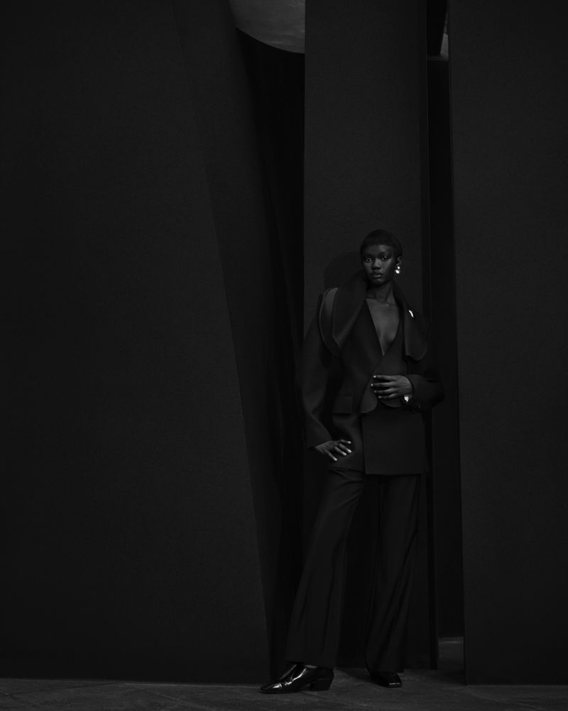 A black-and-white full shot of a Black female fashion model posing in a dark suit with wide-leg pants in a dark, minimalist setting.