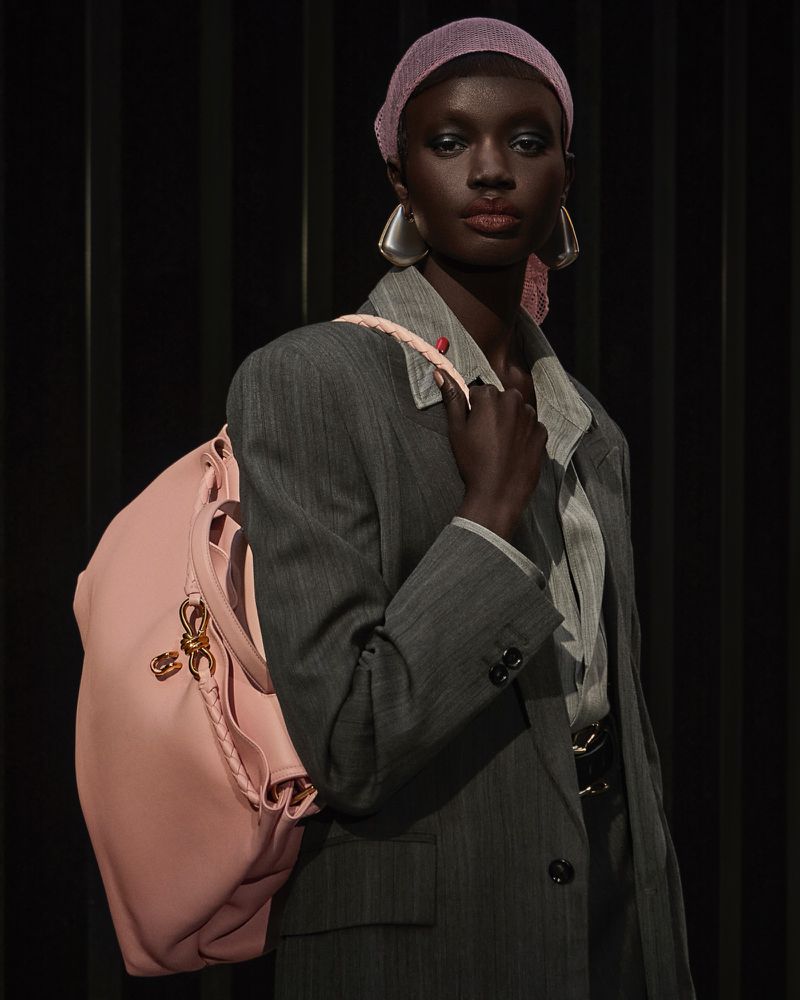 Portrait of a dark-skinned woman with a pink headscarf, gray blazer, silver earrings, and a pink handbag, posing against a dark background.