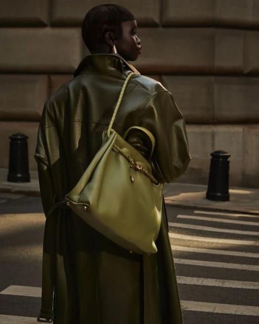 Olive-green leather trench coat and handbag paired with an urban background, worn by a Black female model looking off-camera.