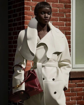 Model Aker Ajak wearing a structured white coat and holding a burgundy handbag outdoors against brick